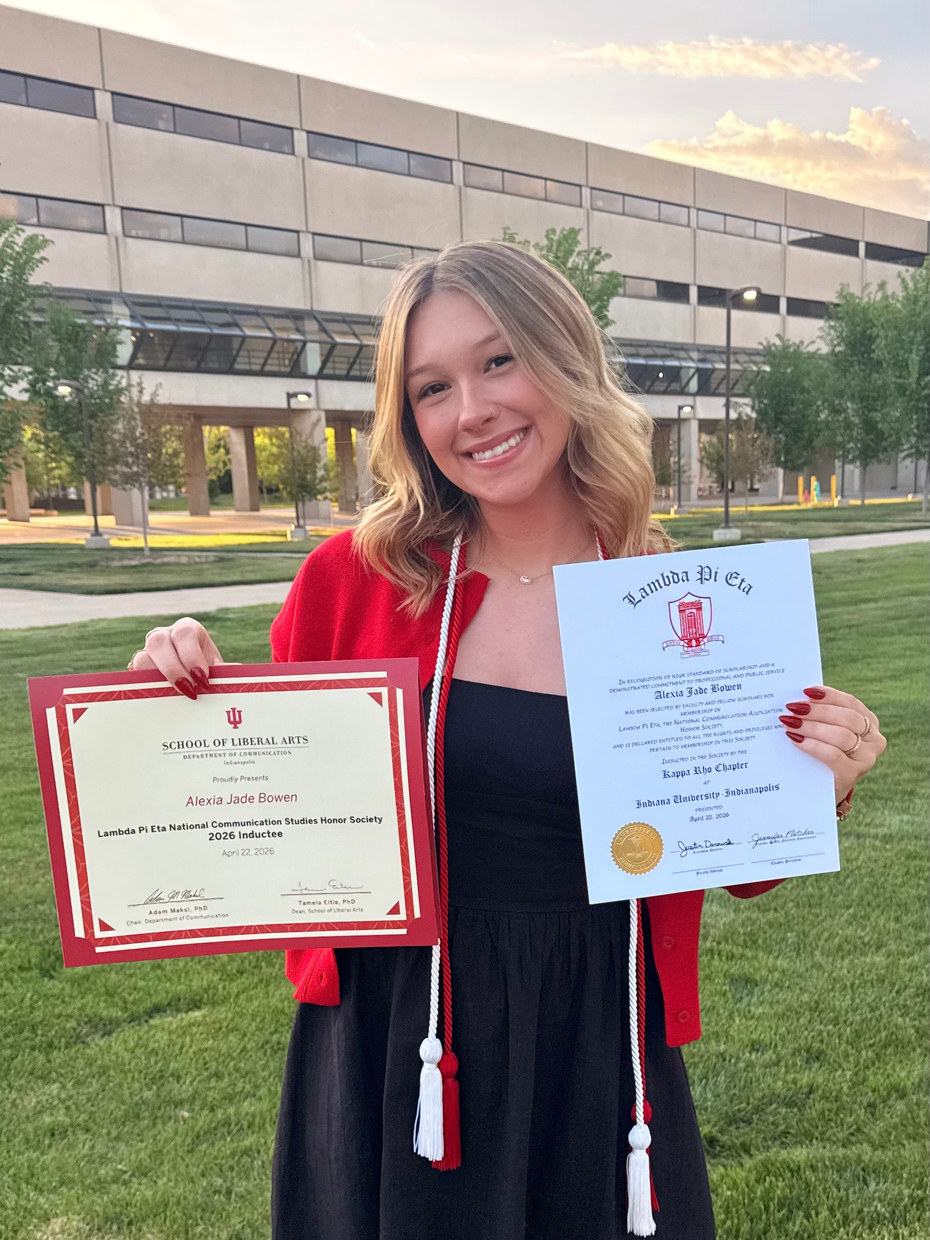 Alexia is holding two certificates for being inducted into the Lambda Pi Eta, posing for a picture on campus.